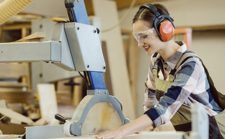 „Klartext „Betriebsarzt überwacht den Einsatz von Schutzkleidung und sicheren Arbeitspraktiken an einer Bandsäge in einer Schreinerei zur Förderung des Arbeitsschutzes.“ „. - © Kzenon – stock.adobe.com