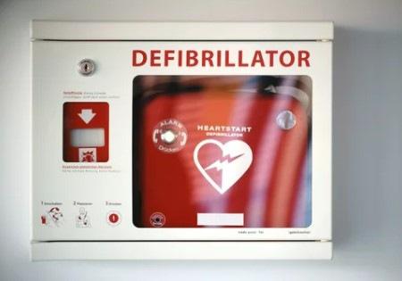 „Wandinstallierter Defibrillator in einer weißen Box mit roter Beschilderung und Anweisungen zur Förderung der Sicherheit am Arbeitsplatz und zur Unterstützung präventiver Gesundheitsmaßnahmen. - © ginton – stock.adobe.com