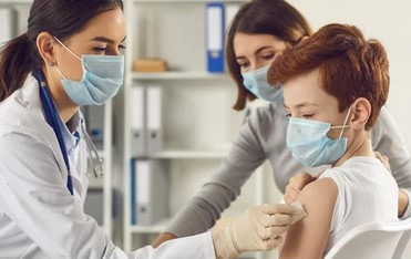 Alt-Text: „Ein Gesundheitsmitarbeiter führt eine Impfung an einem jungen Jungen durch, während eine Frau in der Nähe sitzt; alle tragen Masken. Das Bild verdeutlicht präventive Maßnahmen im Arbeitsschutz und betont die Bedeutung von Impfungen für den Gesundheitsschutz am Arbeitsplatz. - © Studio Romantic – stock.adobe.com