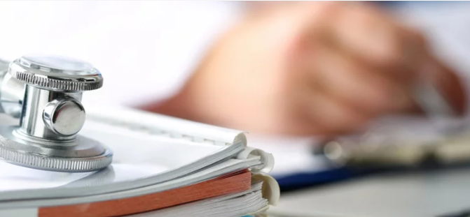 „Ein Stethoskop ruht auf einer Sammlung von Fachbüchern zur Arbeitsmedizin, während im Hintergrund eine Hand sorgfältig Notizen zu präventiven Maßnahmen und arbeitsschutzrelevanten Leitlinien verfasst. - © megaflopp – stock.adobe.com