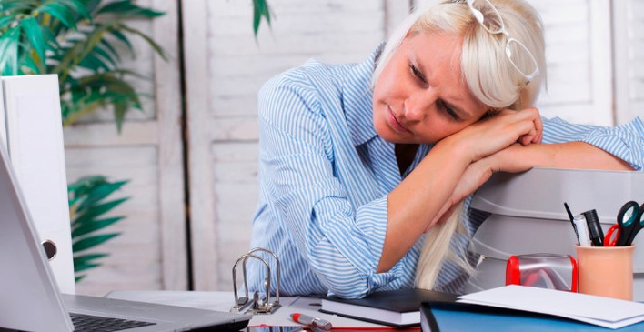 Arbeitsmedizinerin in blauer Bluse zeigt Erschöpfungszeichen am unorganisierten Schreibtisch mit Laptop und Büromaterialien, symbolisiert die Notwendigkeit von präventiven Maßnahmen und ergonomischen Arbeitsplatzlösungen im Arbeitsschutz. - © absolutimages – stock.adobe.com
