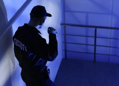 Sicherheitsbeamter in Uniform, der mit einer Taschenlampe ein schwach beleuchtetes Treppenhaus überprüft. - © New Africa – stock.adobe.com