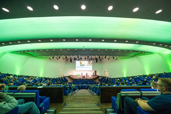 Großes Auditorium mit blauen Sitzen, einer Bühne und einer grün beleuchteten Decke; man hört den Rednern zu. - © Basi / Bernd Lauter